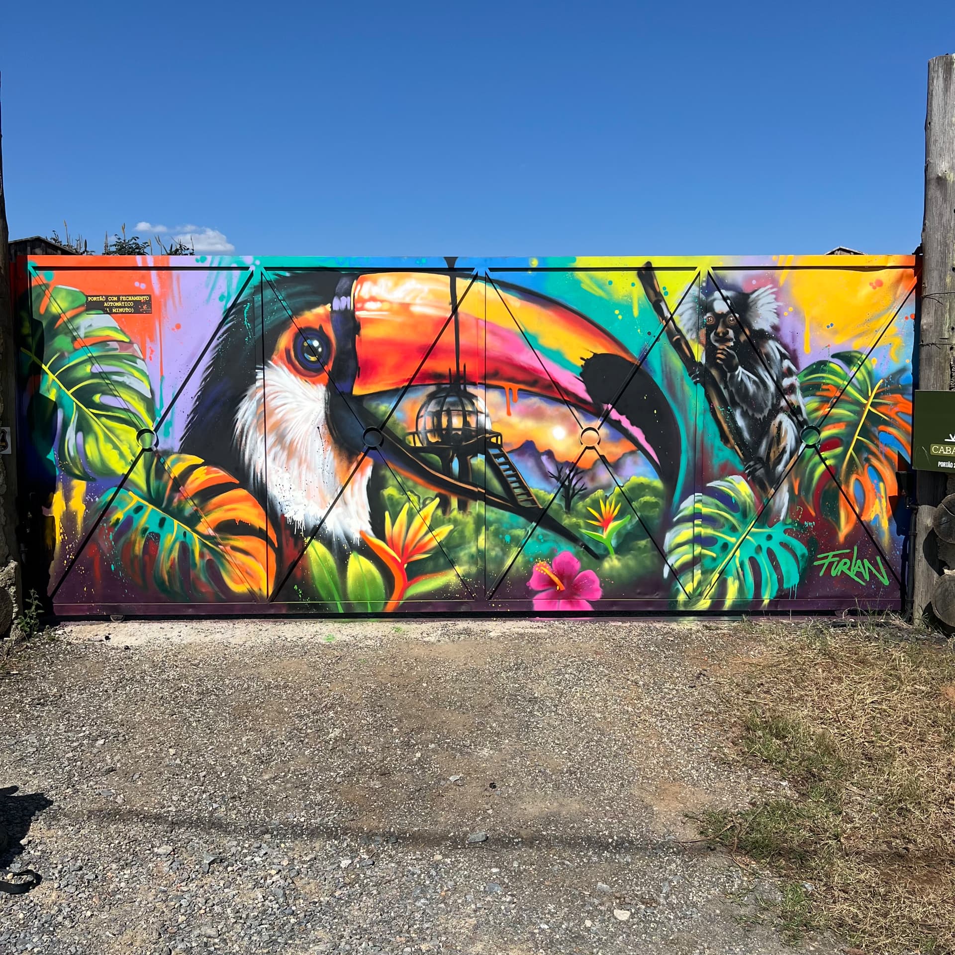 Mural colorido em portão com tucano, primata, folhagens e flores, fotografado ao ar livre